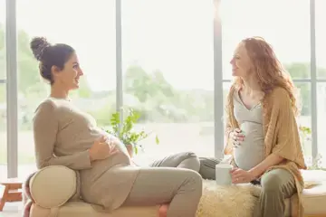 Zwei schwangere Frauen sitzen auf einer gepolsterten Bank vor einem Fenster und unterhalten sich. Die Sonne scheint, beide lächeln und sehen entspannt aus.