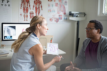 Ein Mann sitzt in einem Sprechzimmer einer Arztpraxis. Ihm gegenüber sitzt eine Ärztin und hört ihm aufmerksam zu. An der Wand hängen Poster, die das Innere des Menschen zeigen, auf dem Computerbildschirm der Ärztin ist ein Röntgenbild zu sehen.