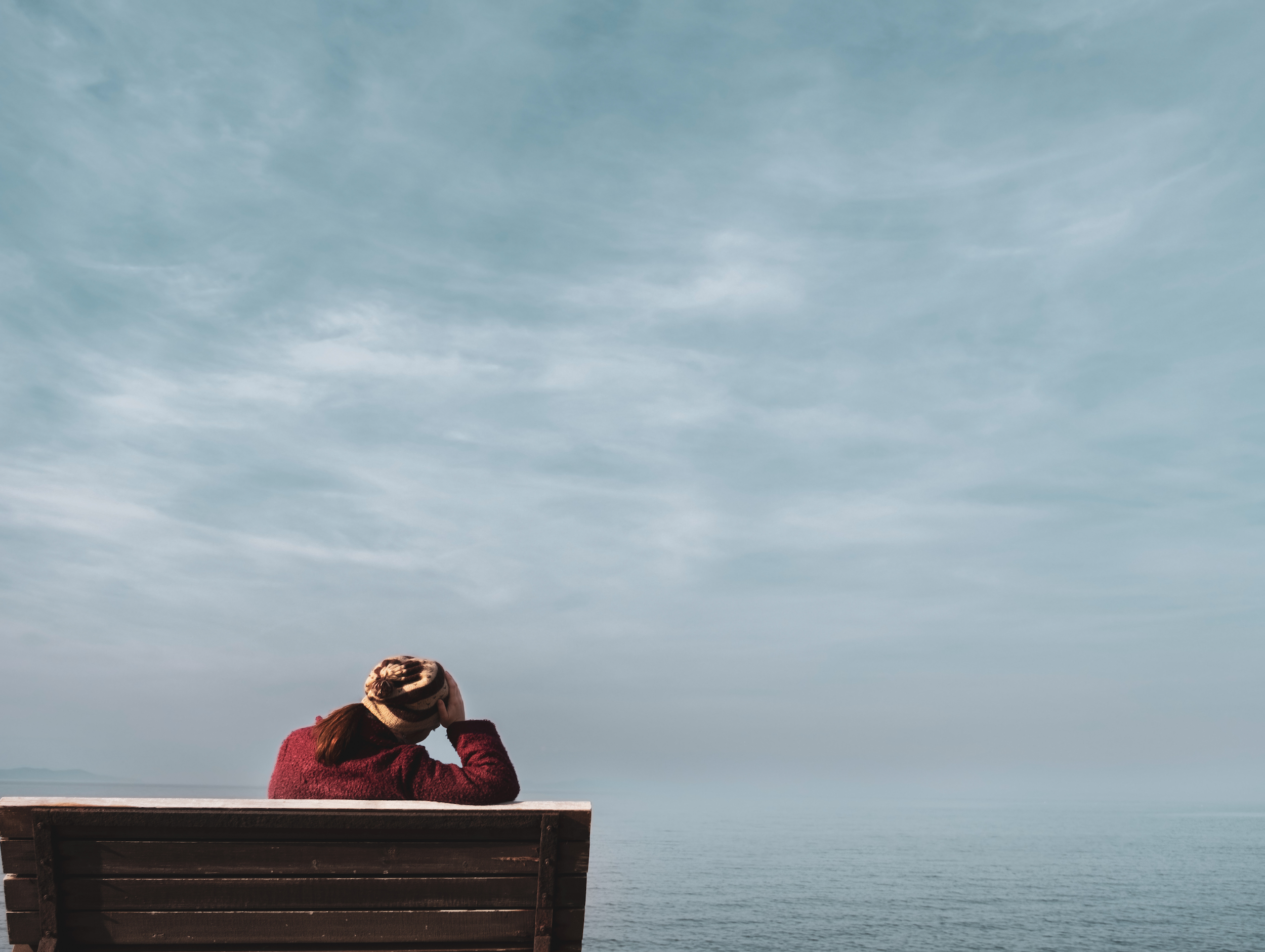 BZgA Organspende Blog Identität Spender 1 Eine Frau sitzt auf einer Bank. Sie hat ihren rechten Arm auf der Rücklehne abgestützt und hält ihren Kopf. Sie ist von hinten zu sehen. Im Hintergrund sieht man das Meer und den Himmel.