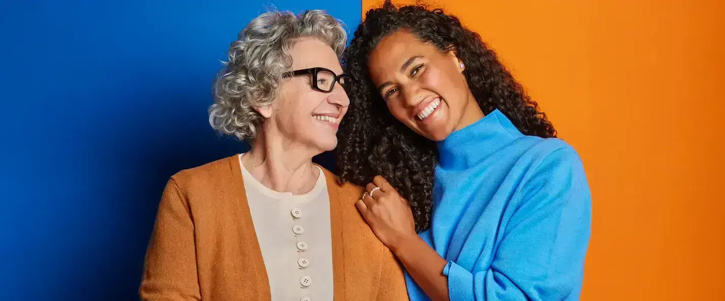 Zu sehen sind eine ältere Frau mit orangenem Cardigan auf blauem Hintergrund und eine jüngere Frau mit einem blauen Pullover auf orangenem Hintergrund. Beide Frauen lächeln und stehen vertraut aneinander.