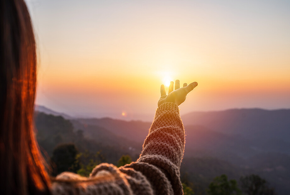 Eine Frau wird von hinten fotografiert, wie sie ihren Arm austreckt und der aufgehenden Sonne entgegenstreckt. Dabei steht sie auf einem Berg, das Tal und Bergketten im Hintergrund sind erkennbar.