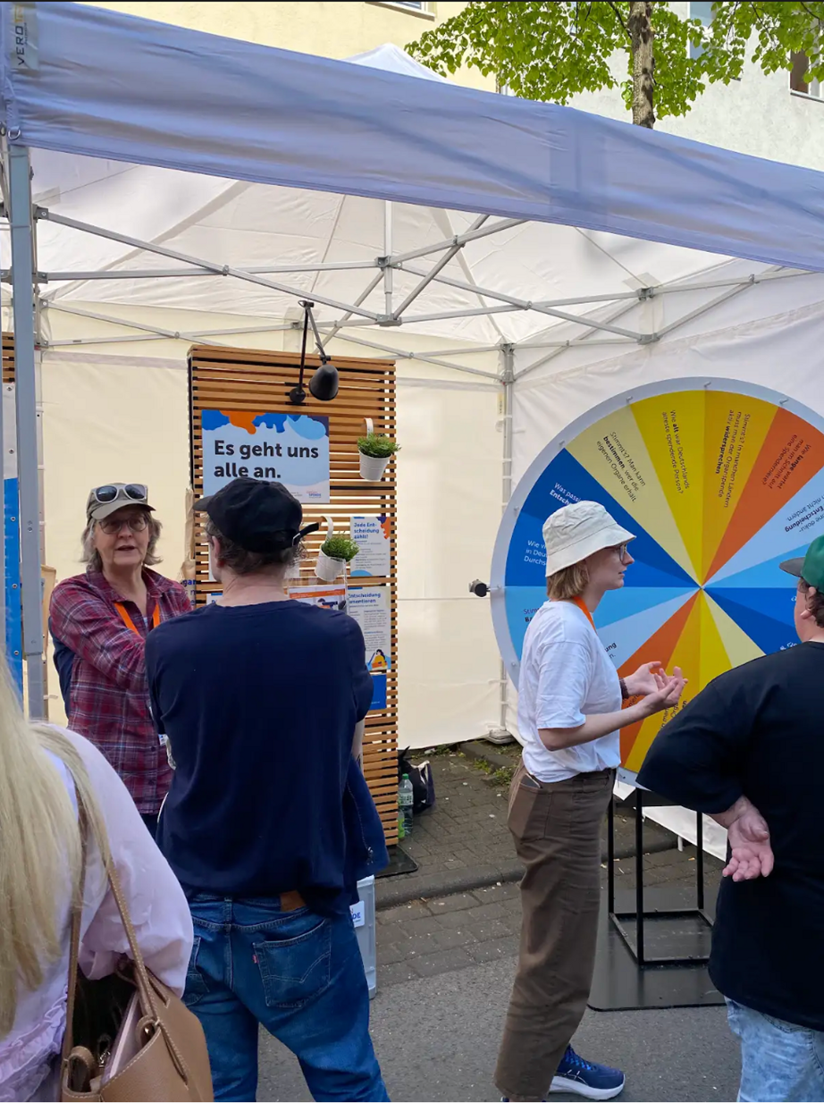 Das Bild zeigt den Stand des BIÖG Organspende auf der c/o pop 2025, mit Menschen im Gespräch und dem Glücksrad.